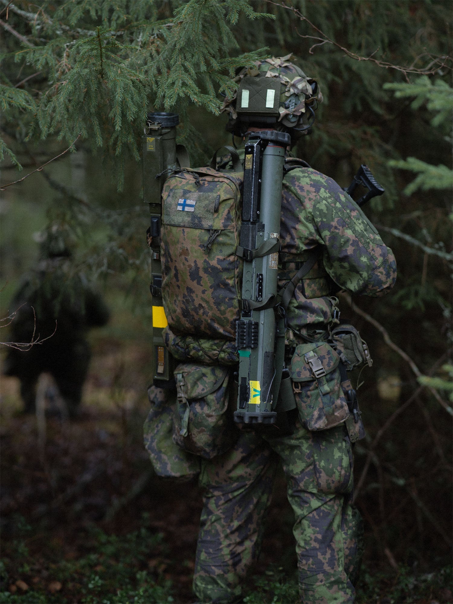 Person in camouflage military gear with a Savotta Niukka 20L backpack and NVD counterweight pouch attached to the helmet, in a forest setting