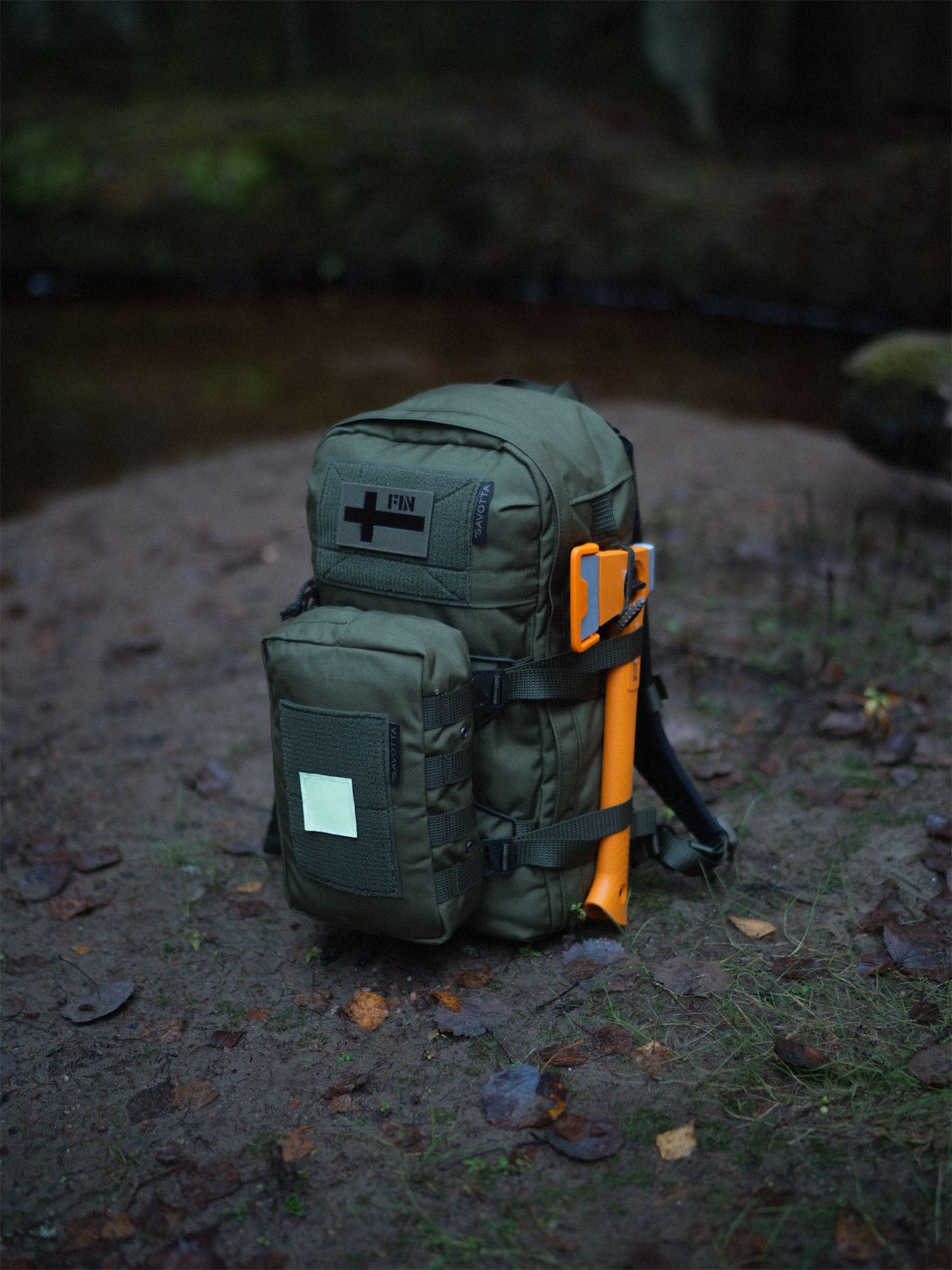 Green Savotta Niukka 20L backpack with Savotta Suur-kukkaro attached to the front and an orange axe to the side, on a forest floor
