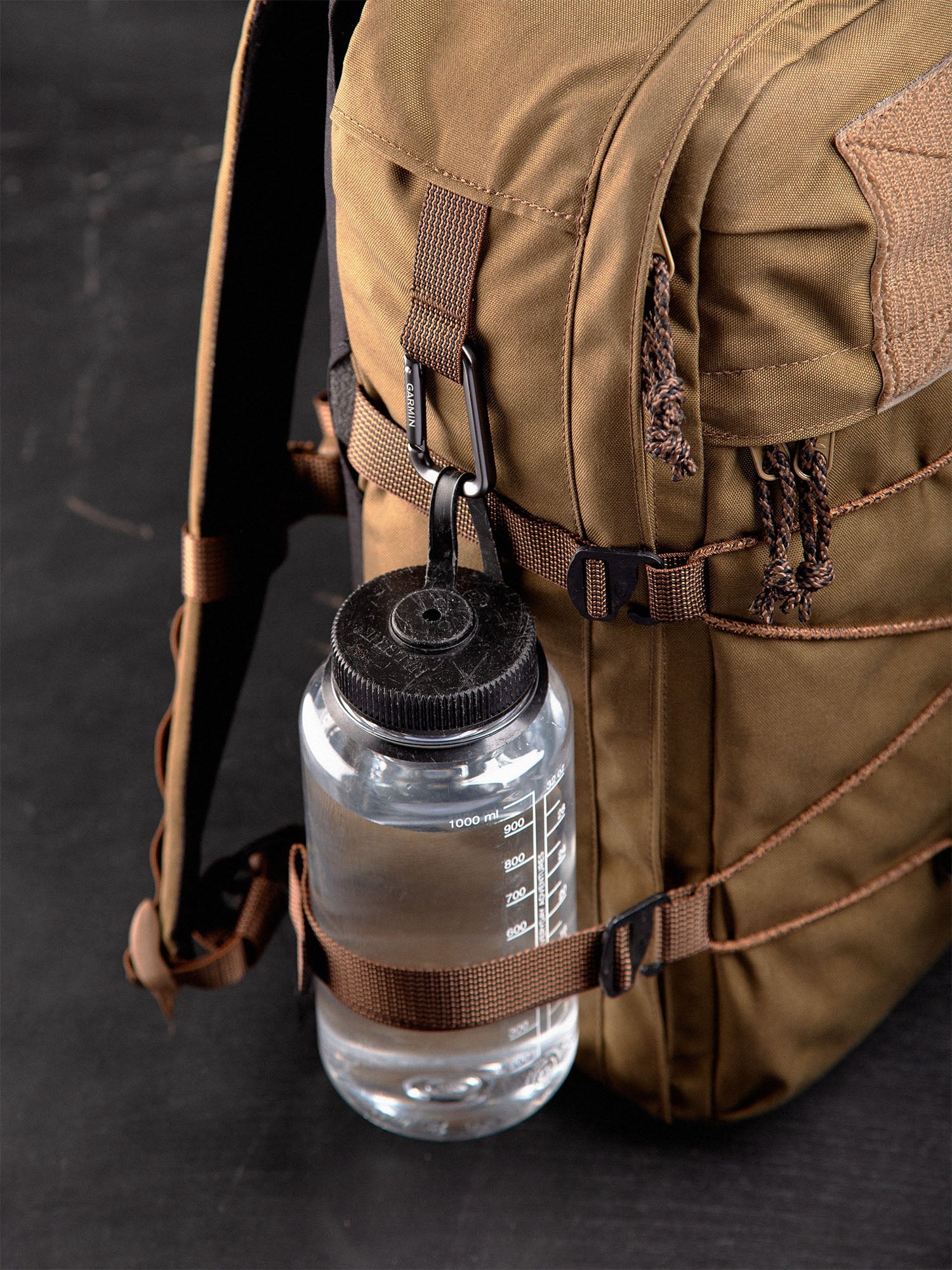 Brown Niukka 20L backpack with a Nalgene water bottle attached to the side with a carabine and the side compression strap on a dark background