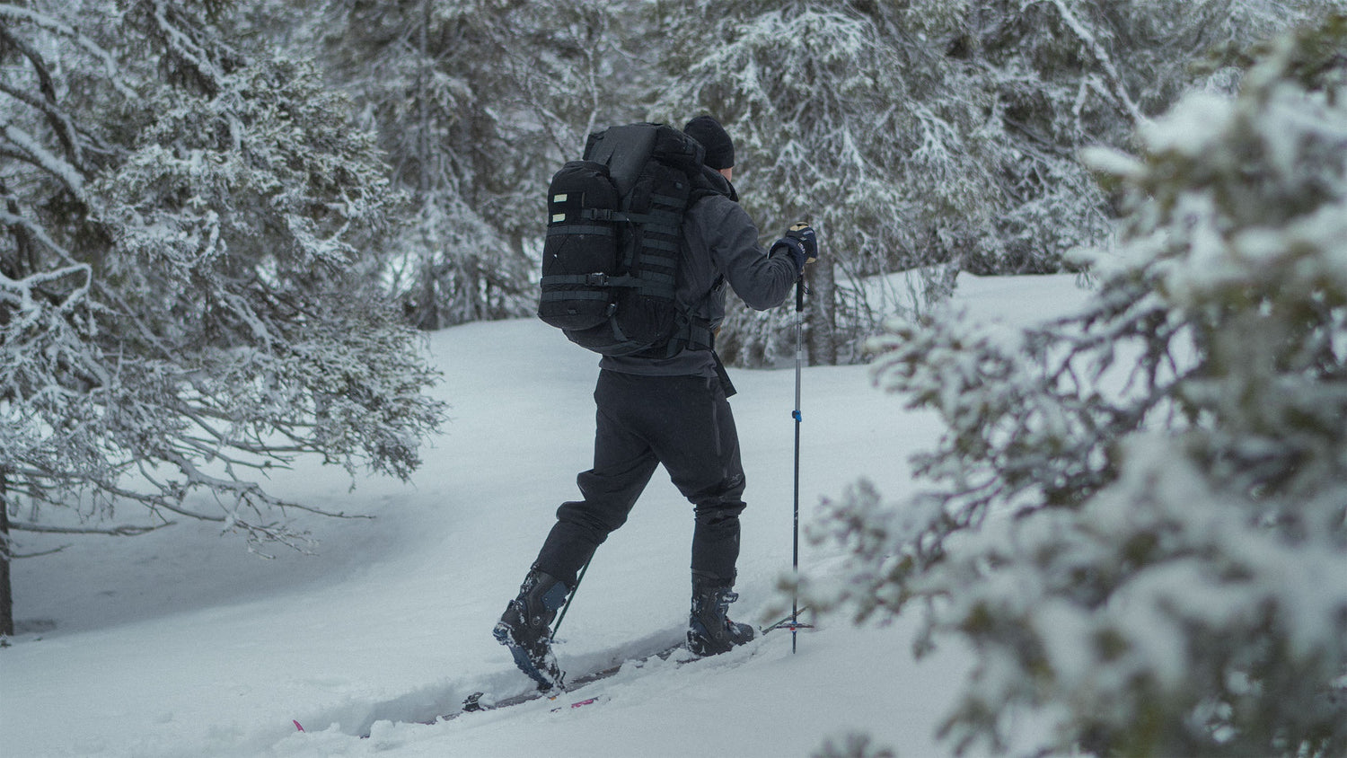 Person in black outdoor clothing cross-country skiing through a snowy forest with a Savotta Kantamus backpack and Savotta Hatka bag attached