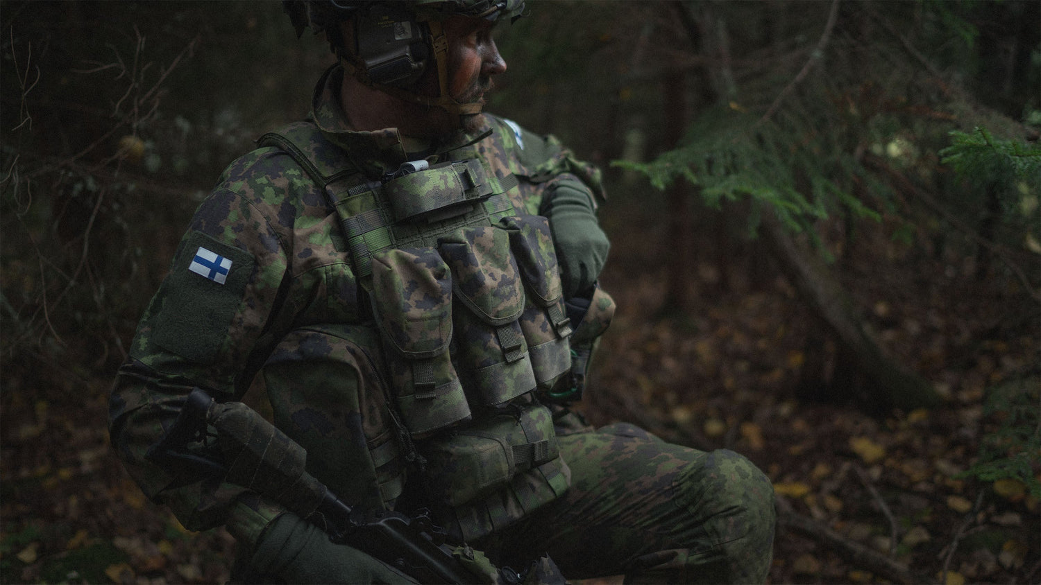 Person in military camouflage gear with a Savotta Varjelus plate carrier. There are multiple pouches attached to the plate carrier. A dark forest background