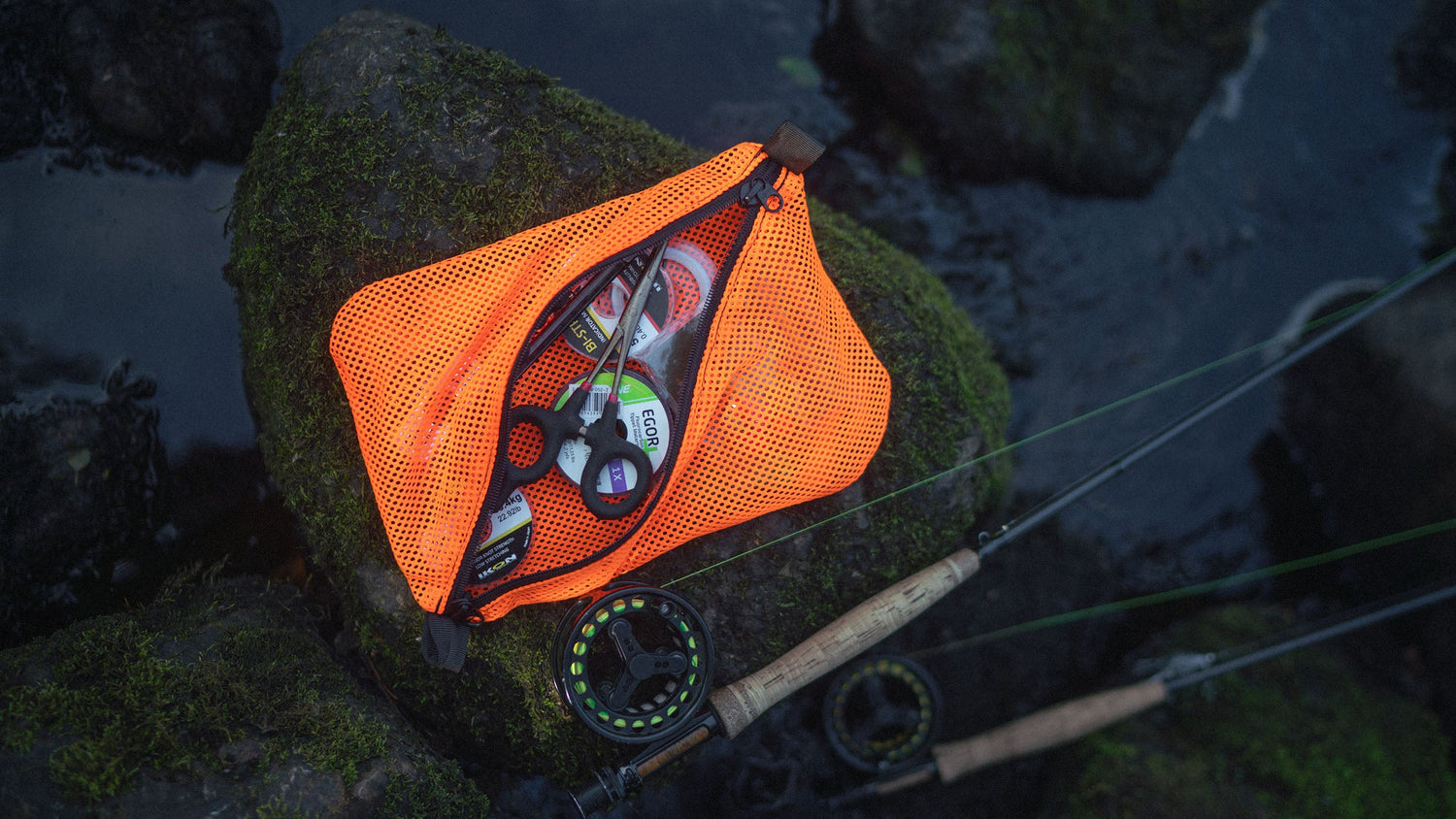 Orange Savotta trinket pouch on a rock surrounded by water with fishing equipment nearby