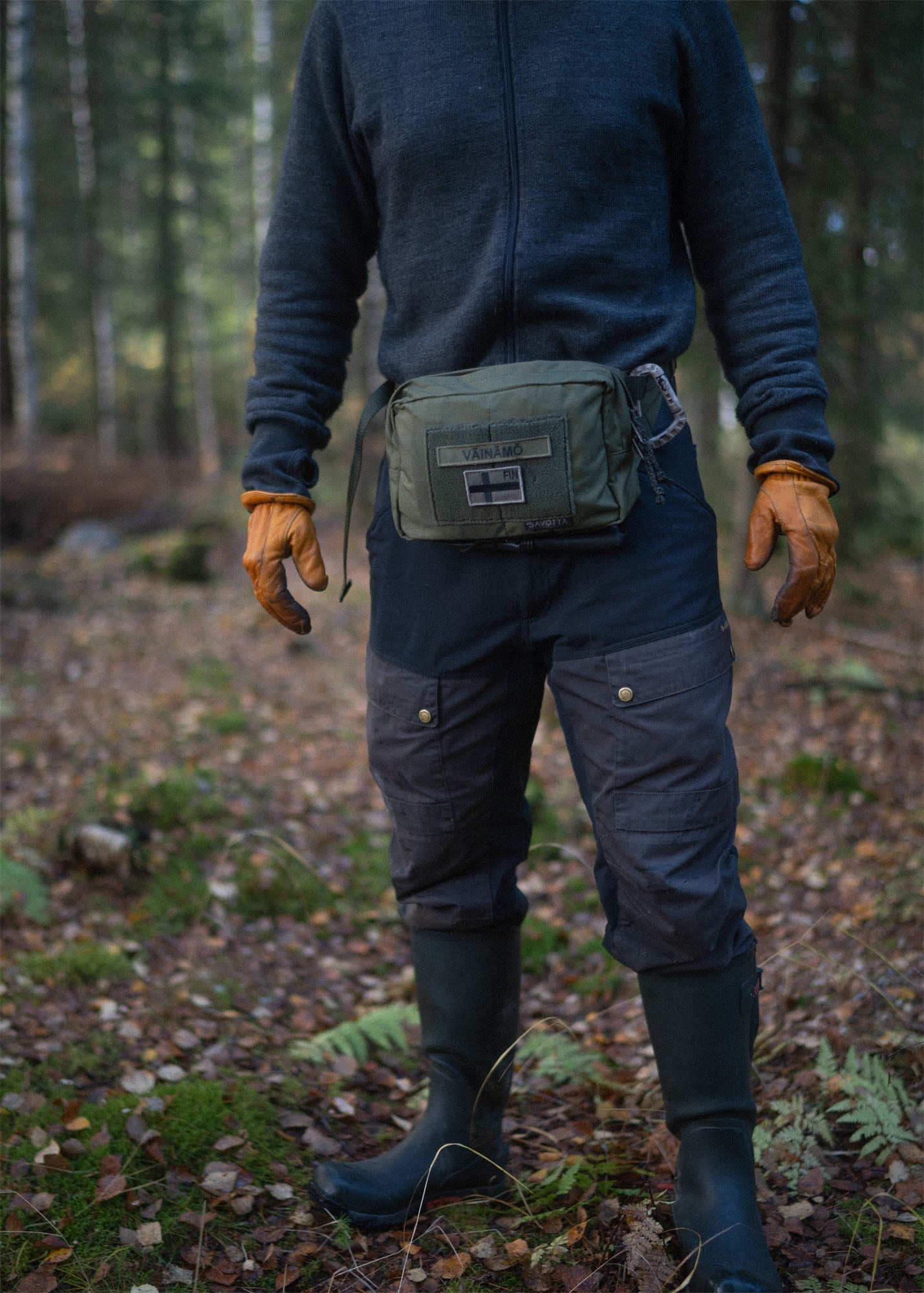 Person in a forest wearing a Savotta Suur-kukkaro hip pack, blue jacket, and rubber boots.