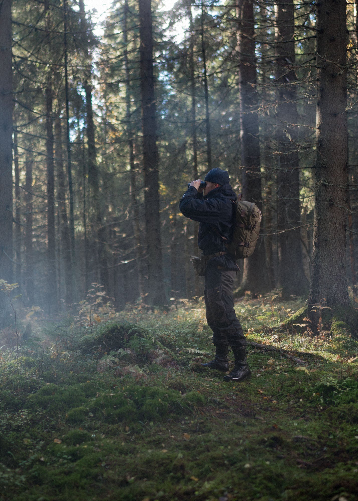 Person in outdoor clothing with a Savotta Niukka 20L backpack and Savotta Suur-Kukkaro hip pack standing in a sunny and foggy forest, holding binoculars.