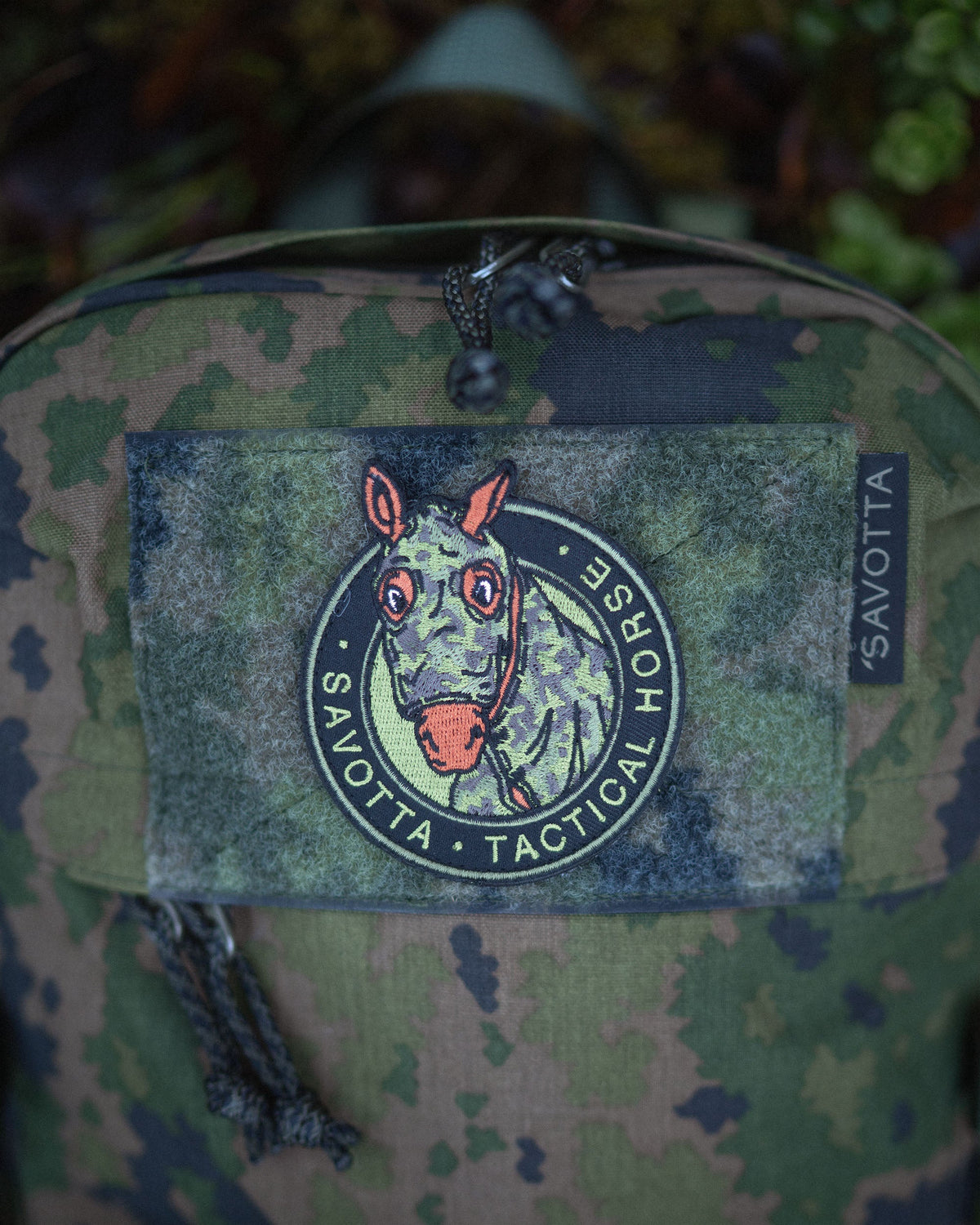 A Savotta Niukka backpack with a horse-themed patch labeled 'Savotta Tactical Horse' on a camouflage background. The horse is wearing camouflage gear.