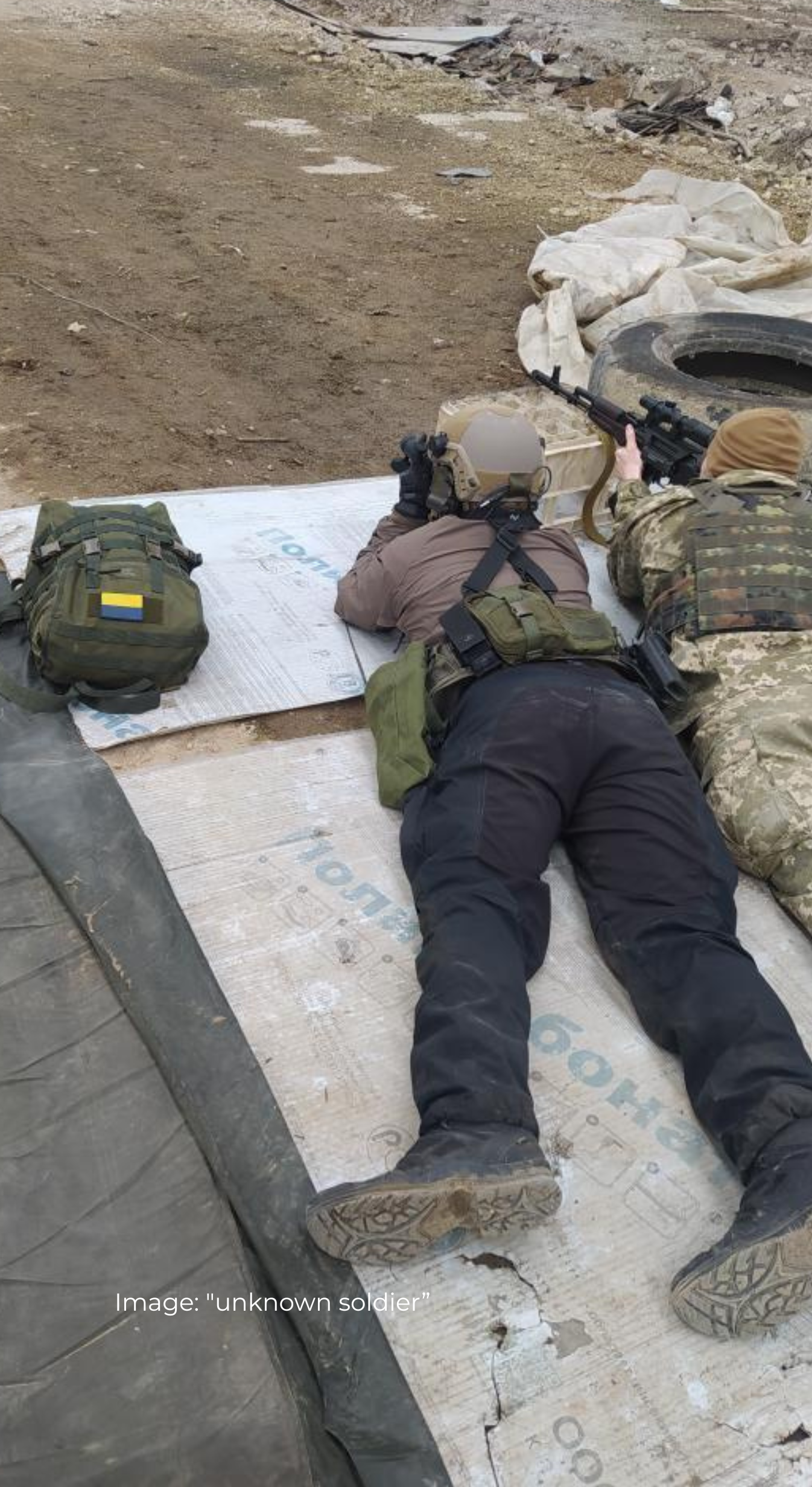Two Ukrainian soldiers in camouflage gear lying on the ground with weapons and Savotta gear, on a gritty ground.