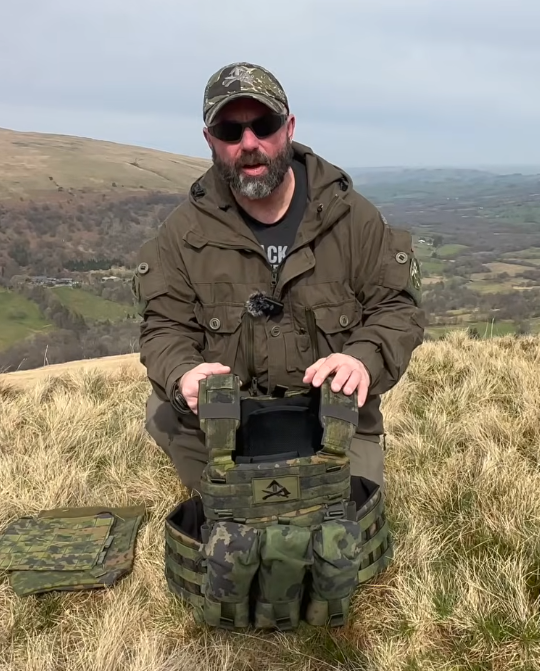 Man holding a Savotta Varjelus plate carrier on top of a grassy hill.
