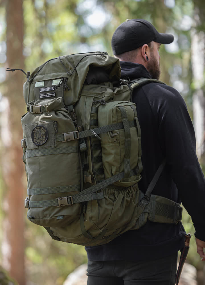 Person wearing a green Kantamus 60L backpack with Hatka 12L attached to the front and side 4L pouches  in a forest setting