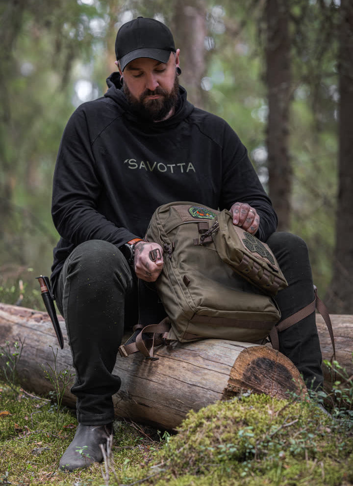 Man sitting on a log in a forest, wearing a black 'Savotta' hoodie and holding a brown Savotta Niukka 20L backpack.