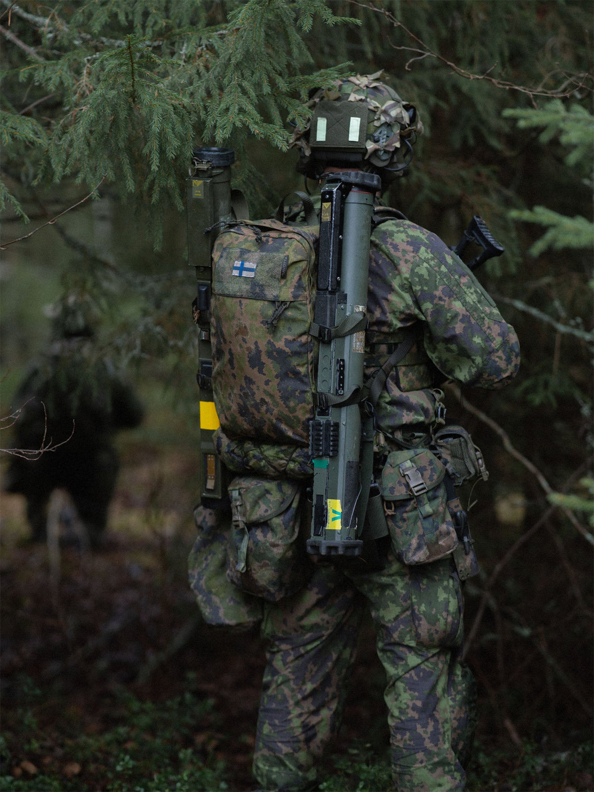 Person in camouflage military gear with a Savotta Niukka 20L backpack and NVD counterweight pouch attached to the helmet, in a forest setting