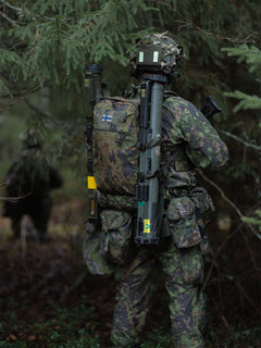 Person in camouflage military gear with a Savotta Niukka 20L backpack and NVD counterweight pouch attached to the helmet, in a forest setting