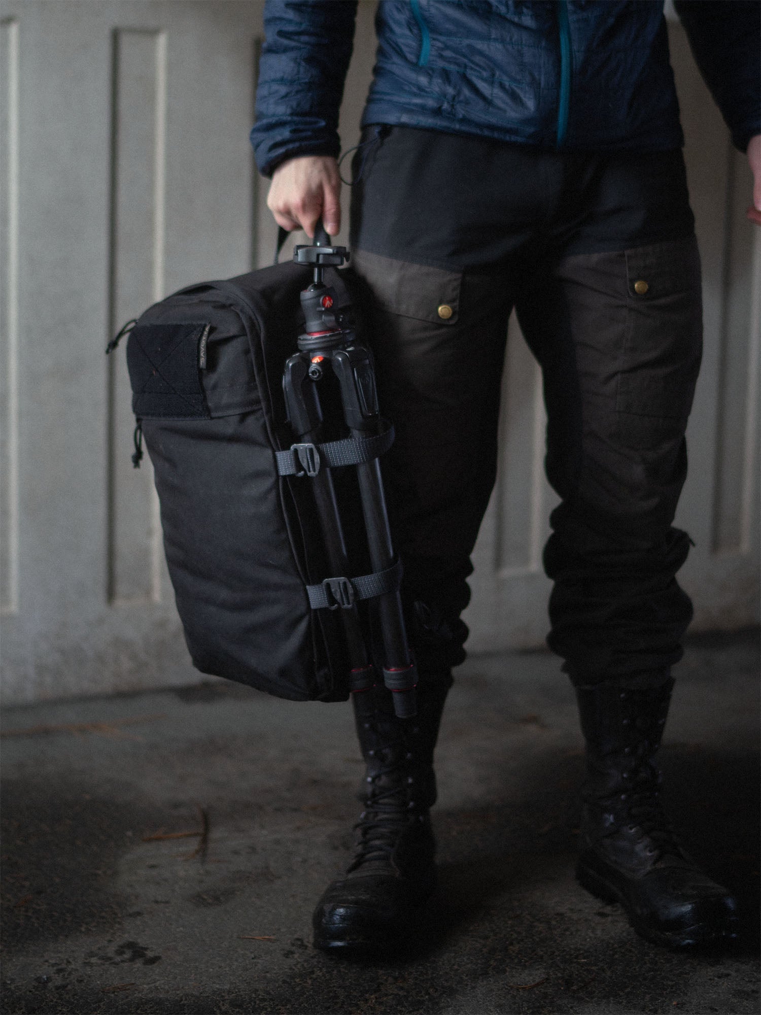 Person holding a black Savotta Niukka 20L backpack with a camera tripod attached, wearing dark outdoor clothing and boots.