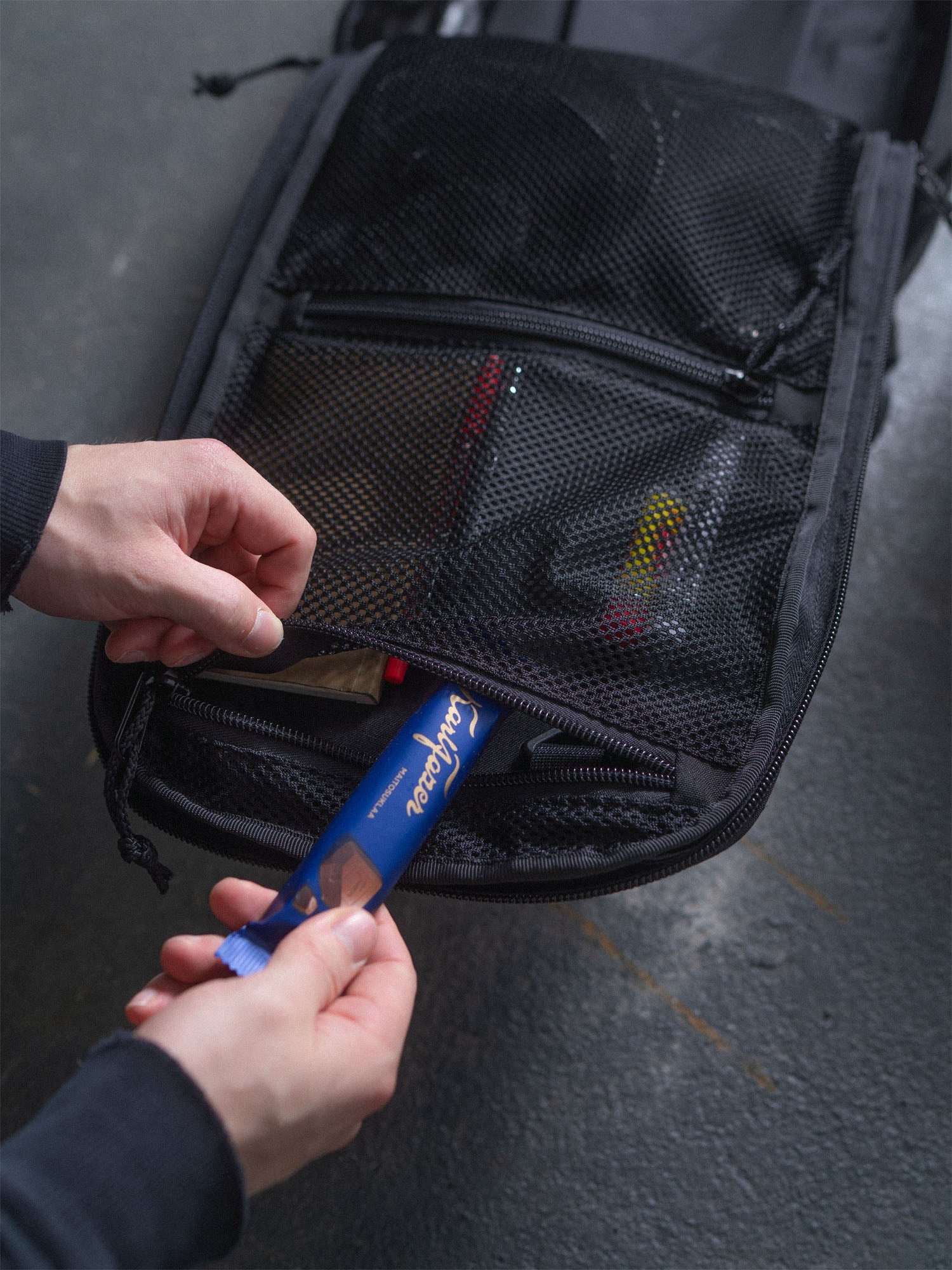 Person putting a chocolate bar into the zipper pouch inside a black Savotta Niukka 20L backpack on a gray surface.