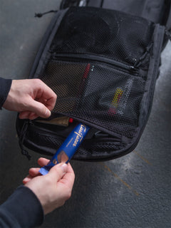 Person putting a chocolate bar into the zipper pouch inside a black Savotta Niukka 20L backpack on a gray surface.