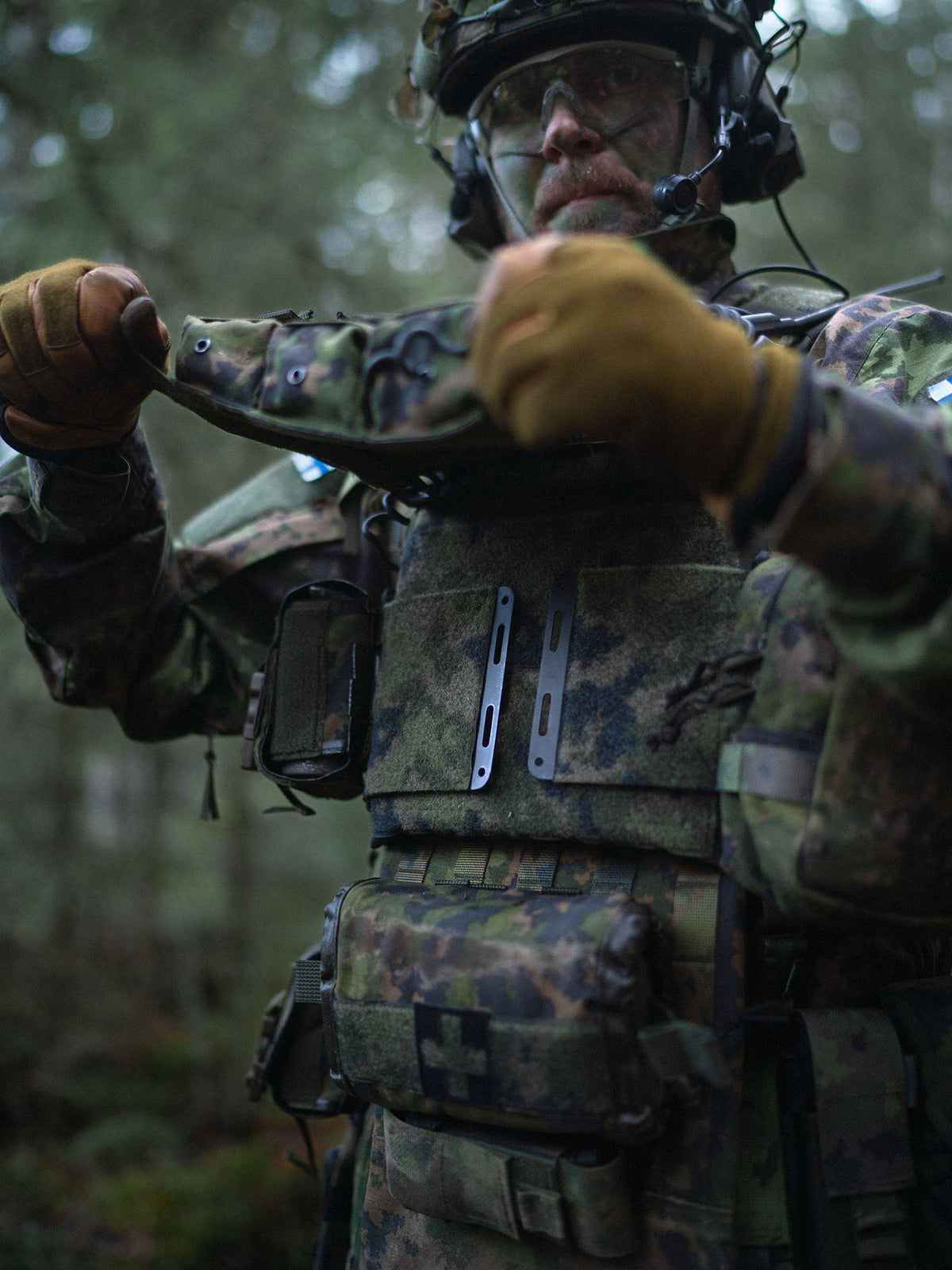 Person in military camouflage gear lifting the Savotta Varjelus 3x2 AR15-AK placard from a Varjelus plate carrier in a forest setting