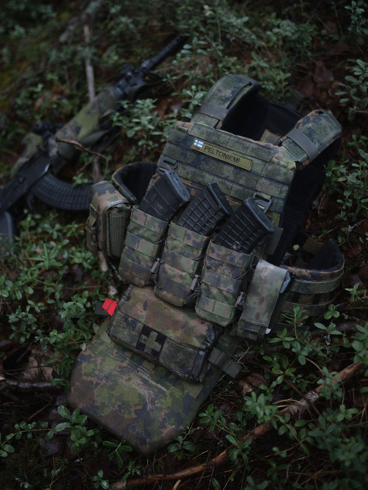 Savotta Varjelus plate carrier with Varjelus groin flap and mag pouches attached, on a forest bed with a rifle in the background