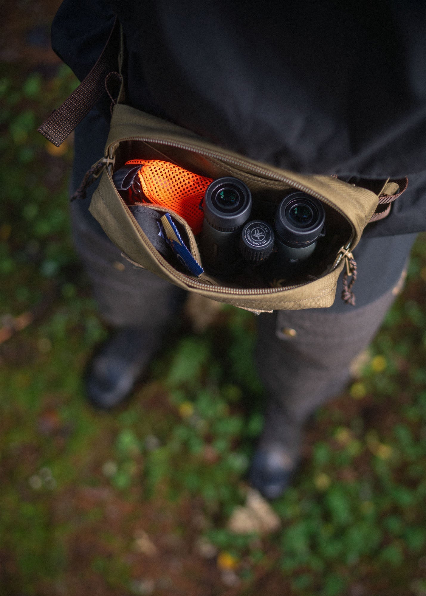An opened Savotta Suur-Kukkaro hip pack on a person in forest, inside the bag there are binoculars, trinket pouch and snacks.