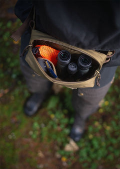 An opened Savotta Suur-Kukkaro hip pack on a person in forest, inside the bag there are binoculars, trinket pouch and snacks.