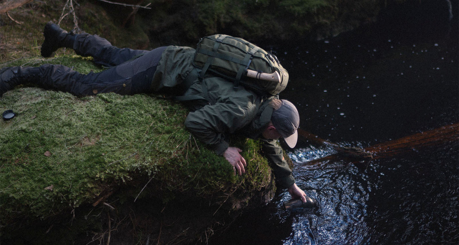 Person in outdoor clothing and a Savotta Kahakka 25L backpack lying on a mossy bank next to a stream and filling his water bottle.