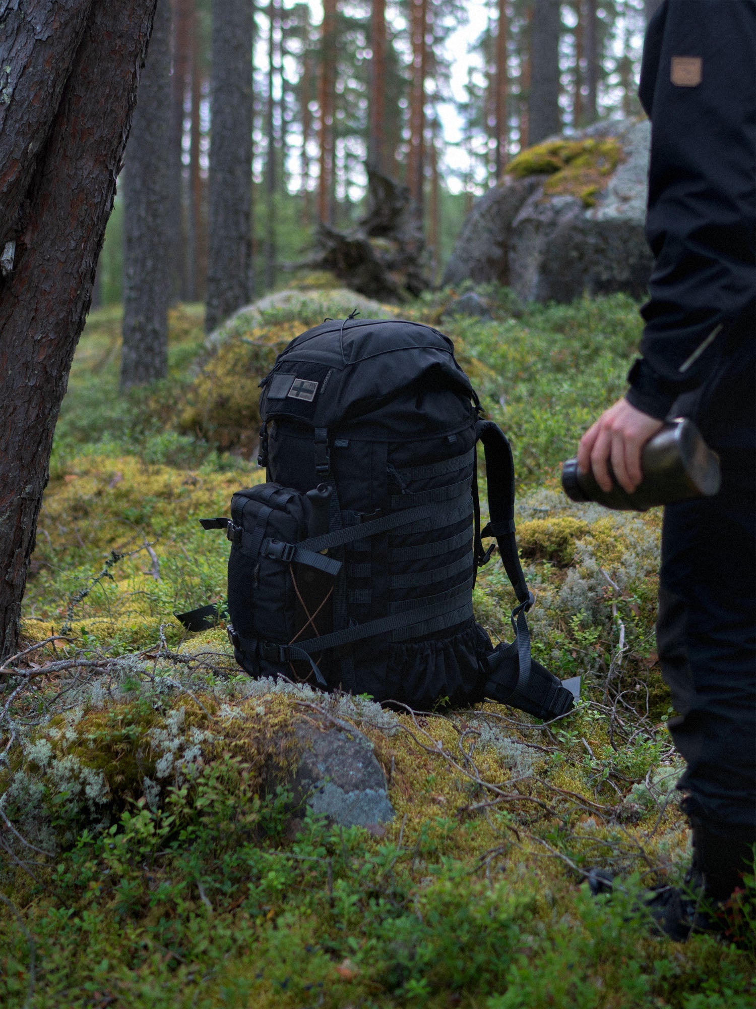 Black Savotta Kantamus 60L with Askare hip pack on a mossy forest floor next to a person holding a bottle.