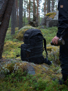 Black Savotta Kantamus 60L with Askare hip pack on a mossy forest floor next to a person holding a bottle.