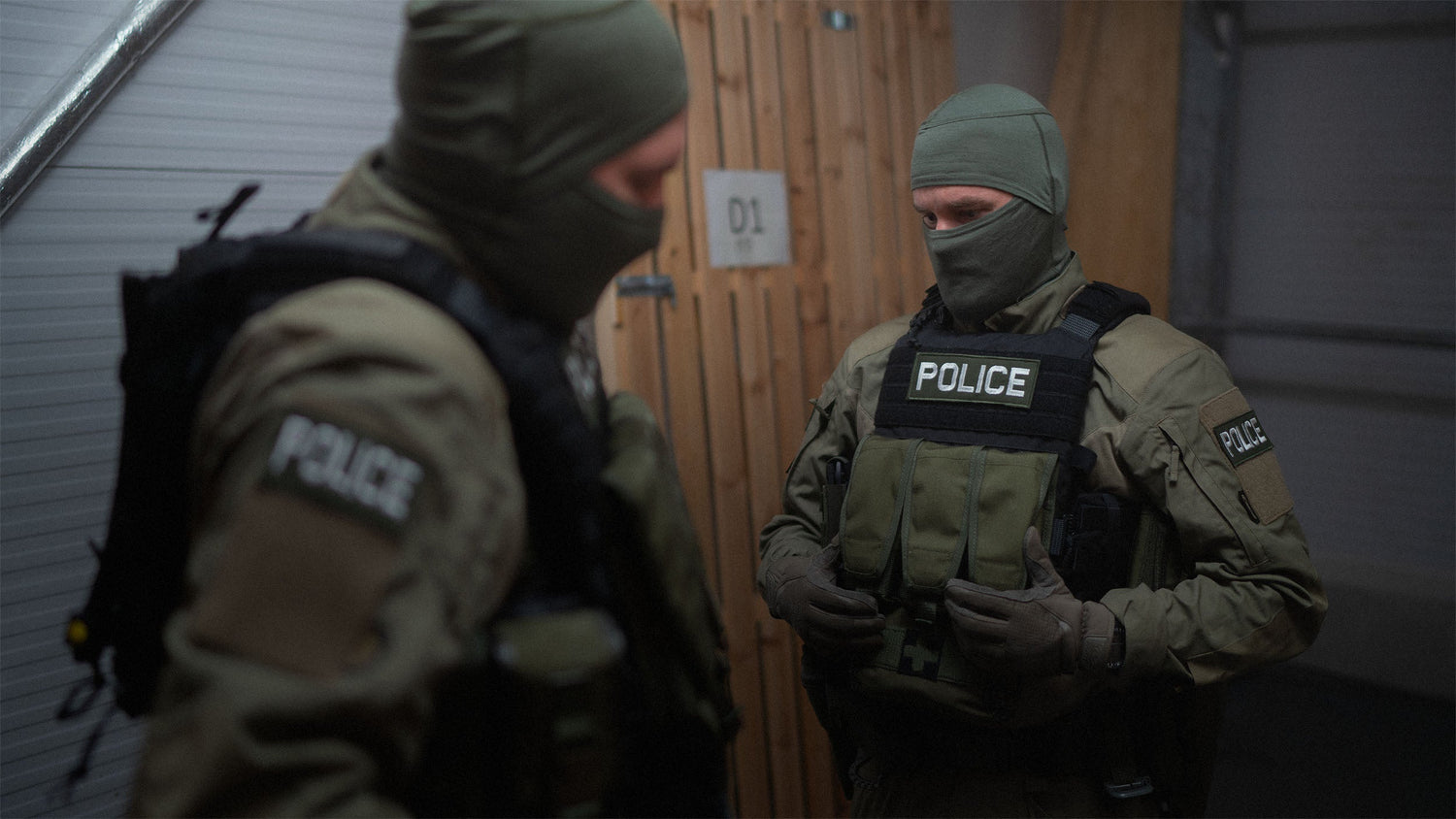 Two police officers in tactical gear standing indoors.