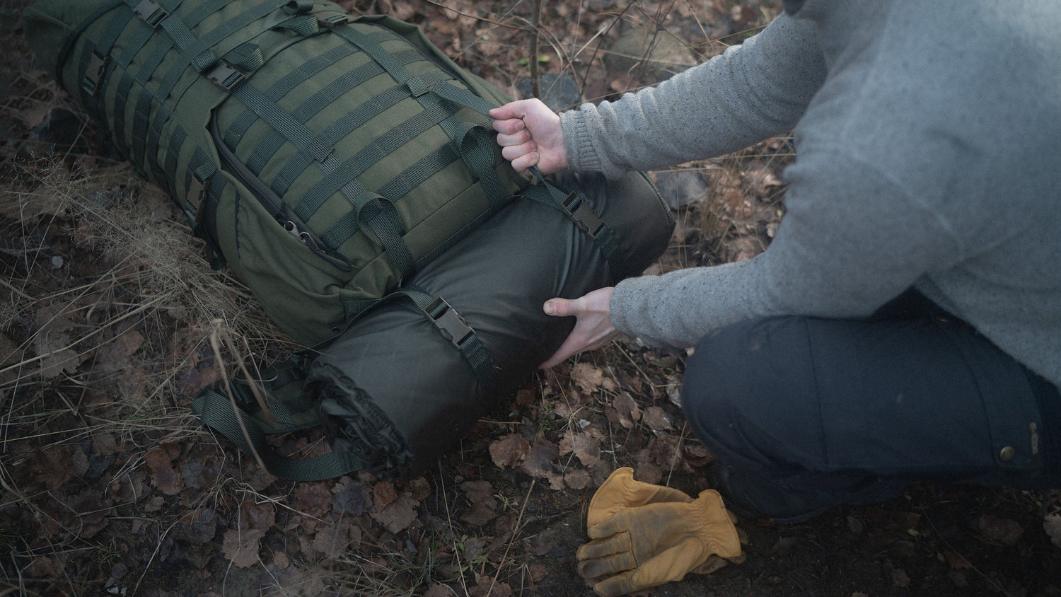 Person attaching a Savotta FDF sleeping pad to a Savotta Jääkäri XL backpack on the ground in a natural setting
