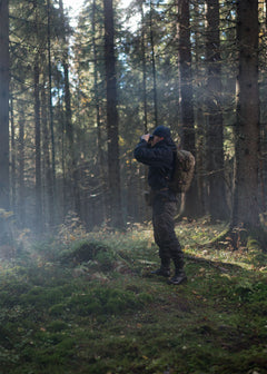Person in outdoor clothing with a Savotta Niukka 20L backpack and Savotta Suur-kukkaro standing in a forest, holding binoculars.