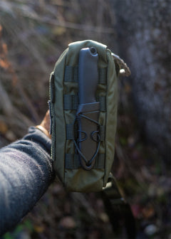 Hand holding a Savotta Suur-Kukkaro hip pack and showing the bottom MOLLE attachment with a blurred natural background
